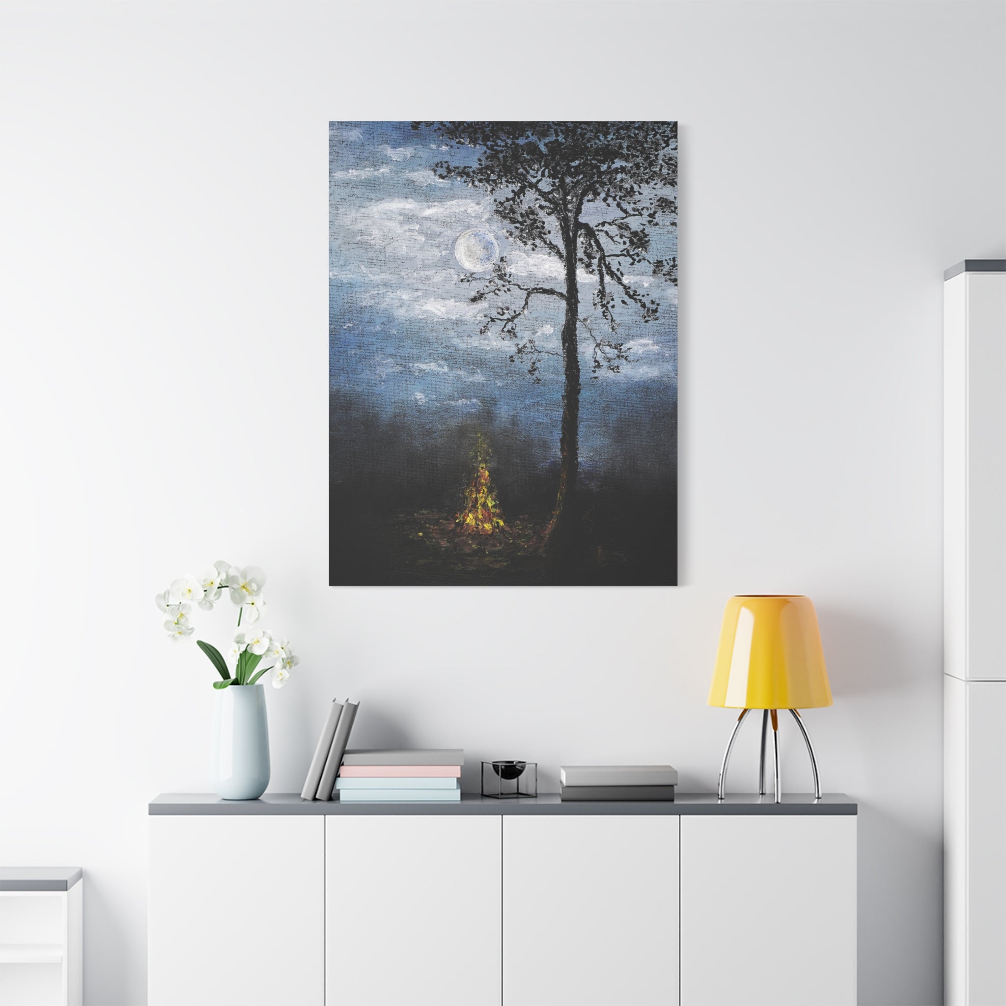 Canvas art printed after an original painting in oil of a forest scene with a campfire on a white wall above a cabinet with books a lamp and decorative objects.