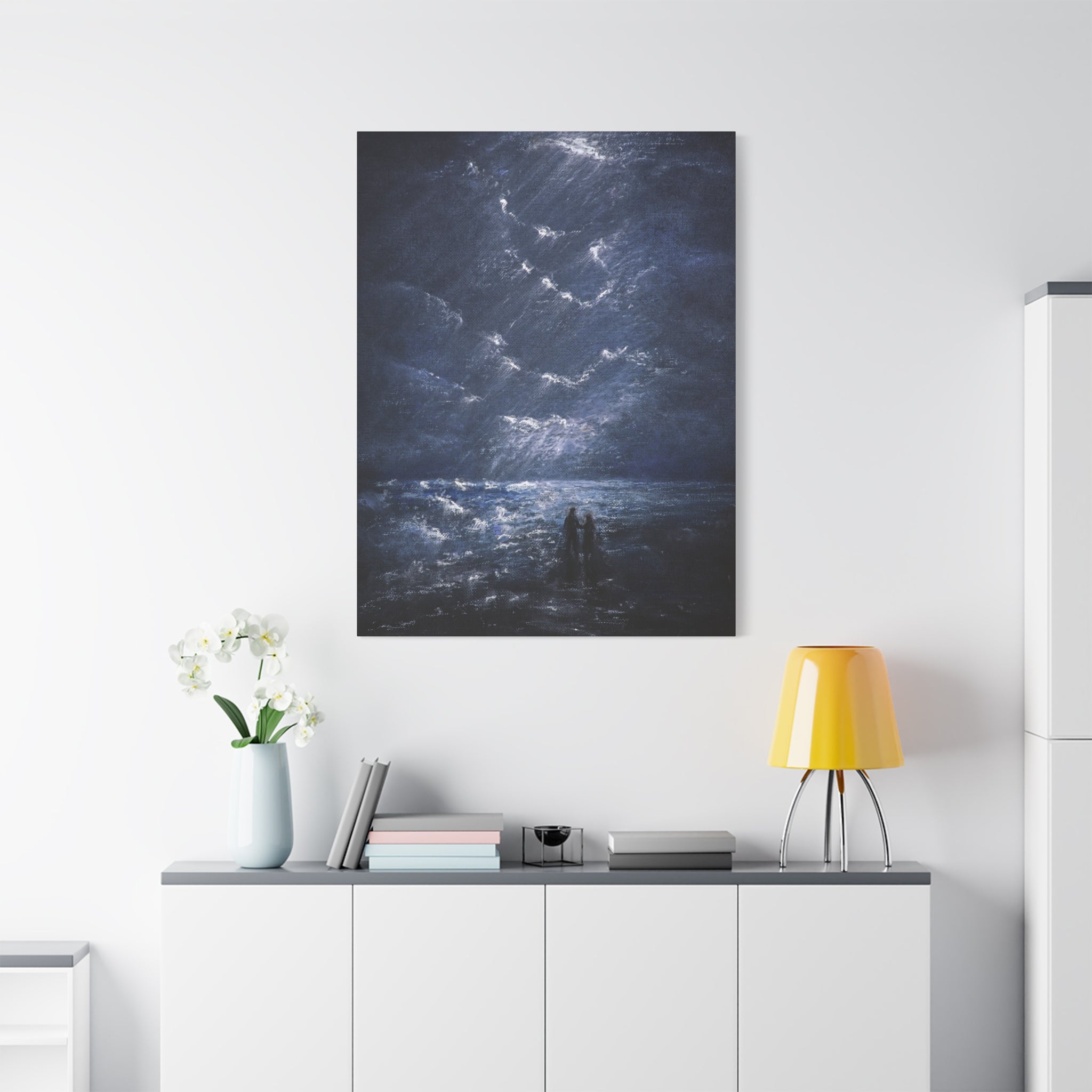 Modern interior with a dark abstract printed matte canvas of a night scene with a couple on the shore, hanging on a white wall above the desk with a yellow lamp, and white cabinet.